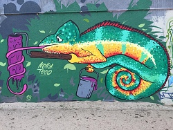 Graffiti an einer Wand, zeigt ein buntes Camäleon mit einem Farbeimer und einer Farbrolle, um die es seine lange Zunge gewickelt hat