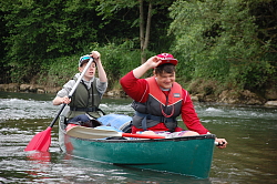 Das Bild zeigt zwei Jungs im Kanu auf dem Wasser. Der hintere paddelt, der vordere fasst sich gerade mit der rechten Hand an seine Baseball-Kappe.