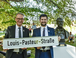 60 Jahre Städtepartnerschaft mit Dole: OB Ibert steht neben Doles Bürgermeister, beide halten ein Straßenschild mit dem Namen Louis-Pasteur-Straße in den Händen. 