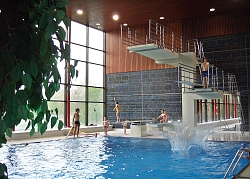 Das Bild zeigt den Fünf-Meter-Sprungturm im Hallenbad und das becken darunter mit einigen Badegästen auf dem Turm, im Wasser und am Rand.
