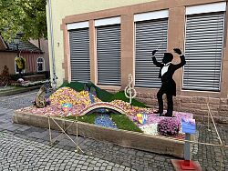 Blütenwagen mit Figur eines Dirigenten und einer Brücke über einen Bach