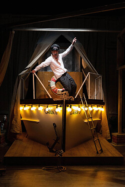 Zu sehen ist ein Schauspieler der in gelbem Licht angeleuchtet wird. Er posiert alleine auf einem Holzschiff 