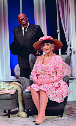 Eine ältere Dame im Kleid sitzt auf einem Sessel, ein älterer Herr im Anzug steht hinter ihr und spricht zu ihr