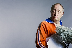 Mann mit orangener Sweatjacke und Puschelmikrofon vor grauem Hintergrund.