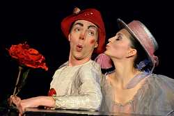 Clown mit rotem Hut und roter Kunstblume macht ein verwundertes Gesicht, während ihm eine Frau mit Tüllkleid und Hut mit gespitzten Lippen einen Kuss auf die Wange geben will.