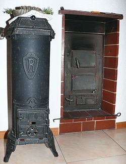 Das Bild zeigt einen alten Guteisernen Kanonenofen, daneben die Befeuerungsklappen für einen Kachelofen.