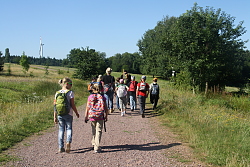 Zu sehen ist eine Gruppe von Kindern mit zwei Betreuern. Sie gehen einen Schotterweg im Wald entlang.