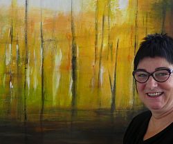 Die Künstlerin Stephanie Hirsmüller vor einem ihrer Bilder. Stephanie Hirsmüller lacht. Sie trägt eine schwarze Brille, ein schwarzes Shirt und kurze dunkle Haare.