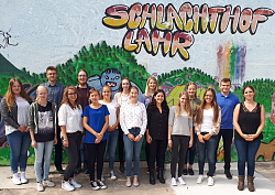 15 neue Auszubildende beginnen ihre Berufskarriere bei der Stadt Lahr. Die Auszubildenden stehen vor einer Wand mit einem Graffiti und der gesprühten Schrift "Schlachthof Lahr". 