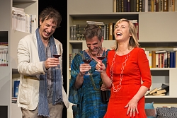 Drei Schauspieler, zwei Männer und eine Frau, stehen, modern gekleidet, zusammen und trinken ein Glas Wein.