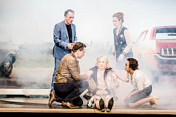 Zu sehen ist eine Szene aus der Komödie „Bella Figura“ von Yasmina Reza mit dem Ensemble des Euro-Studio Landgraf. Auf der Bühne sind fünf Personen: Eine blonde Frau sitzt, die Beine nach vorn gestreckt, auf dem Boden, links von ihr kniet ein Mann, rechts eine Frau. Beide sprechen die Dame in der Mitte an. Dahinter stehen ein weiterer Mann und eine Frau. Im Hintergrund sind rechts und links teilweise Autos zu sehen. Das Bühnenbild liegt im Nebel, wie von Autoabgasen. Die Szene stellt einen Autounfall dar.