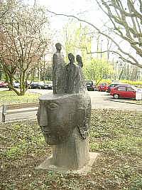 Steinskulptur in Form eines Kopfes, der über den Augen abgeschnitten ist. Auf dieser Kopffläche sind drei Figuren ab der Bauchregion angebracht. Wiese und Bäume im Hintergrund 