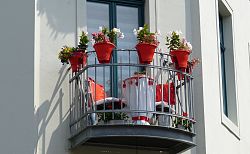 Das Bild zeigt einen kleinen Balkon mit Metallgeländer, darauf stehen ein kleiner runder Tisch und zwei Stühle sowie mehrere mit Blumen bepflanzte Gefäße. Am oberen Rand des Balkongeländers sind fünf große bunt bepflanzte Blumentöpfe angebracht. Dahinter eine zweiflüglige Balkontür. 