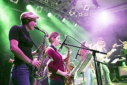 Auf einer Bühne steht im Scheinwerferlicht eine Band und musiziert. Im Vordergrund zwei junge Saxophonistinnen.
