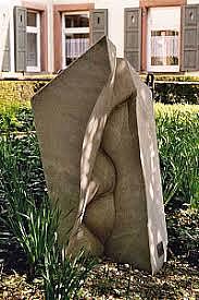 Steinskulptur, die aussieht wie der Innenbereich von verschränkten Fingern. Wiese und Sträucher im Hintergrund