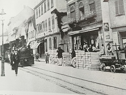 Auf dem historischen schwarz-weiß Foto von 1902 fährt das Lahrer Bähnle durch die Stadt. Man sieht Menschen am Rande, die mit ihren Waren auf den ZUg warten.
