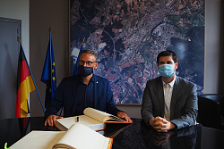 Das Foto zeigt zwei Männer mit Mund-Nasen-Schutz. Sie sitzen an einem Tisch und haben zwei große Bücher vor sich.