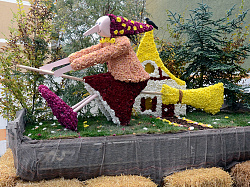 Das Bild zeigt einen bunten Wagen auf dem eine Hexe als Figur mit einem Besen sitzt. Die Figur ist aus Chrysanthemenblüten hergestellt. Das Bild ist sehr bunt. 