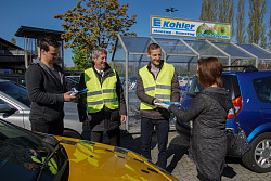 Das Bild zeigt Baubürgermeister Tilmann Petters (2. von links) und Martin Stehr vom Stadtplanungsamt (2. von rechts)in gelben Warnwesten, die auf dem Arena-Parkplatz Brötchentüten, die für den Schulterblick werben, an einen  Autofahrer links, der neben seinem gelben Auto steht, und eine Autofahrerin rechts die gerade zu ihrem blauen Auto geht, verteilen.