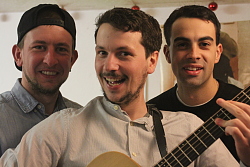 Die drei Musiker stehen nebeneinander. Das Bild zeigt nur die Oberkörper. Dominik Büchele hält in der Mitte eine Gitarre und lächelt keck in die Kamera. Seine zwei Kollegen stehen versetzt dahinter und lächeln ebenfalls. 