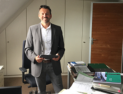 Christian Zanger, Leiter des Rechnungsprüfungsamts der Stadt Lahr, an seinem Arbeitsplatz: Christian Zanger hat dunkles kurzes Haar und einen kurzen Bart, er trägt ein weißes Hemd und ein graues Sakko mit einer grauen Hose. Er steht mit einem iPad in der Hand an seinem Schreibtisch, auf dem viele Unterlagen liegen