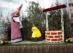 Zu sehen ist ein Chrysanthema Blütenwagen zum Thema Märchen, der eine Szene aus 'Der Froschkönig' darstellt und die Prinzessin und den Frosch mit goldener Krone zeigt sowie einen Brunnen. Alles ist aus Chrysanthemenblüten liebevoll gestaltet.