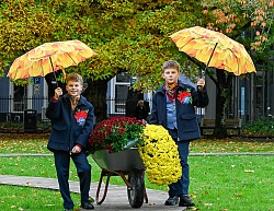 Die Zwillinge mit braunem Haar posieren mit einem orangenem Regenschirm an eine Schubkarre voller Chrysanthemen angelehnt.