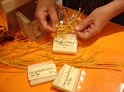 Zwei Hände verpacken ein Stück Seife in Plastikfolie mit einer gelben Schleife aus Bast. Die Seife trägt das Etikett Chrysanthemen-Seife. 