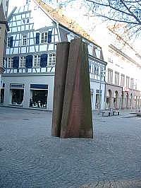Aufrechte geometrische Skulptur auf Kopfsteinpflaster, Hausfassade im Hintergrund