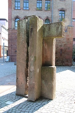Aufrechte Skulptur aus rotem Sandstein auf einem Kopfsteinpflaster-Platz, im Hintergrund eine Hausfassade aus rotem Sandstein und Klinker mit vielen Fenstern.