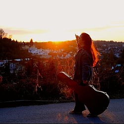Cosima Klenk blickt mit ihrer Gitarre auf den Sonnenuntergang, der sich hinter ihr abspielt. Sie befindet sich auf einer Anhöhe, wodurch die Stadt unter ihr klein aussieht.
