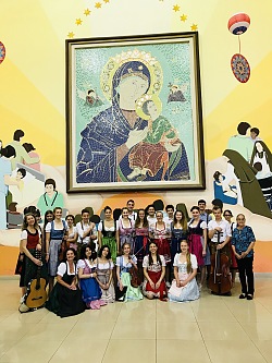 Die Sänger der Kitchen Chicks und die Musiker des Streichquartetts haben sich in verschieden farbigen Dirndl und Tracht vor einem überdimensionalen Mutter Gottes- Bild gruppiert. 