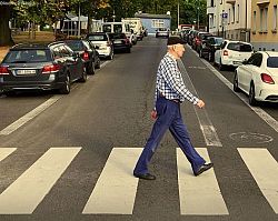 Mann läuft über einen Zebrastreifen, im Hintergrund eine Straße mit seitlich geparkten Autos und Häuserzeile