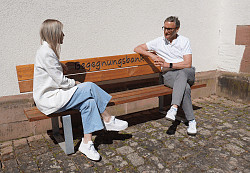 Eine Frau und ein Mann sitzen auf einer Holzbank vor einer Kapelle und sprechen miteinander.
