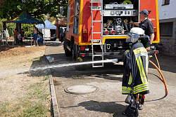 Kind in Feuerwehrschutzkleidung spritzt mit Feuerwehrschlauch Wasser vor einem Feuerwehrfahrzeug auf einem gepflasterten Weg