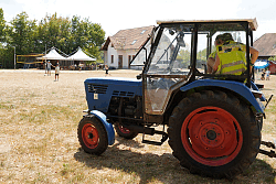 Traktor mit roter Bereifung steht auf einem Feld, im Hintergrund Menschen und Zelte bei sonnigem Wetter