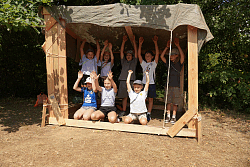 Gruppe von Kindern sitzt und steht in einem selbstgebauten Holzrahmen mit Tuchdach und hebt die Arme
