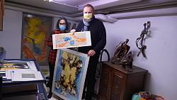 Cindi Ringwald und Guido Schöneboom stehen im Atelier von Ringwald mit Maske und halten zwei Bilder in die Kamera. 