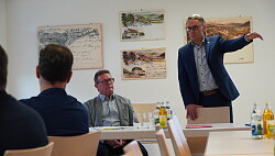 Markus Ibert steht gestikulierend hinter einem Stuh, neben ihn sitzt Ortsvorsther Bühler. Kuhbacherinnen und Kuhbacher hören ihm zu. 