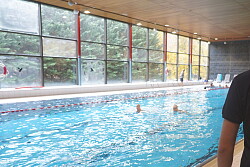 Ein junger Mann steht vor dem Becken im Hallenbad. Er ist von hinten zu sehen und trägt ein blaues T-Shirt mit der Aufschrift Ausbildung. 