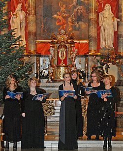 Zu sehen sind sechs Sängerinnen mit schwarzen Kleidern und Liedheften in der Hand im Altarraum einer Kirche. Im Hintergrund ein großes christliches Gemälde, Figuren und prunkvolle Verzierungen. Links hinter den Frauen steht ein Tannenbaum.