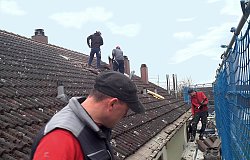 Auf edem Bild sind Dachdecker bei der Arbeit auf einem Ziegeldach zu sehen. Zwei Männerbefinden sich auf einem Gerüst, das am Haus aufgestellt wurde. Am rechten Bildrand ist ein blaues Sicherheitsnetz zu sehen.Auf dem Ziegeldach sind zwei weitere Männer bei der Arbeit.