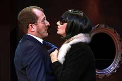 Mann in blauem Anzug mit blauer Krawatte wird an eben dieser von einer Frau gepackt. Sie hat kinnlange schwarze Haare, eine runde schwarze Sonnenbrille und einen schwarzen Mantel mit grauem Fellkragen.