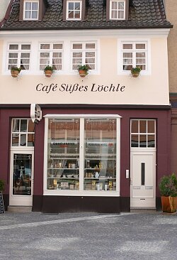 Fassade eines Cafés mit Schriftzug &#39;Café Süßes Löchle&#39;, großen Schaufenstern und drei Blumenkästen unter den oberen Fenstern.