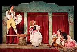 Bühnenbild mit zwei Männern und zwei Frauen. Alle tragen altertümliche Kleidung, die Männer Pumphosen mit weißen Hemden und aufwendig verzierten Westen, die Frauen mehrschichtige und aufwendige Kleider. Roter Samtvorhang im Hintergrund.