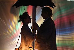 Zwei Personen, von denen nur der Schatten zu sehen ist, halten gemeinsam einen Schirm. Hintergrund ist abstrakt bunt gefärbt.