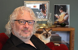 Portrait Ralf H. Dorweiler mit Brille und gewellten grauen Haaren, Vollbart. Im Hintergrund sitzt eine Katze auf der Sofalehne.