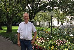 Dr. Michael Dutschke steht im Innenhof am Rathaus 1 in Lahr. Hinter ihm ein kleiner Park mit einem Springbrunnen und bunten Blumen. Er hat graues kurzes Haar, das er zurückgekämmt trägt. Dutschke hat ein weißes Hemnd und eine blaue Jeans an.