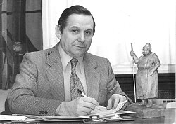 schwarz/weiß-Fotografie Brucker im Anzug mit Kravatte am Schreibtisch mit einem Stift in der Hand. 