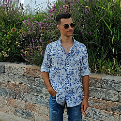 Der junge Mann trägt ein lockeres Hemd und eine Sonnenbrille. Er blickt gelassen nach links, hinter ihm befindet sich eine Mauer, auf der sich Pflanzen und Blumen ranken.